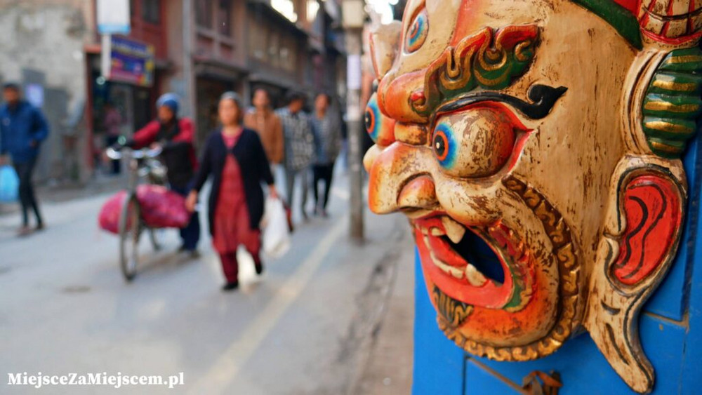 Zdjęcie gościa wydarzenia. Maska drewniana na drzwiach. W tle ulica i spacerujący ludzie.
