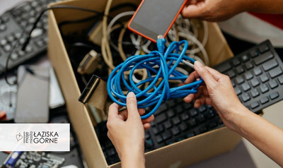 Osoba trzyma niebieski kabel sieciowy nad pudełkiem pełnym zużytego sprzętu elektronicznego i przewodów. W tle widoczne są klawiatury komputerowe i telefon w kartonowym pudle.