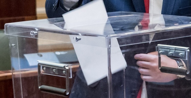 Grafika przedstawiająca wrzucanie głosu do szklanej skrzyni wyborczej