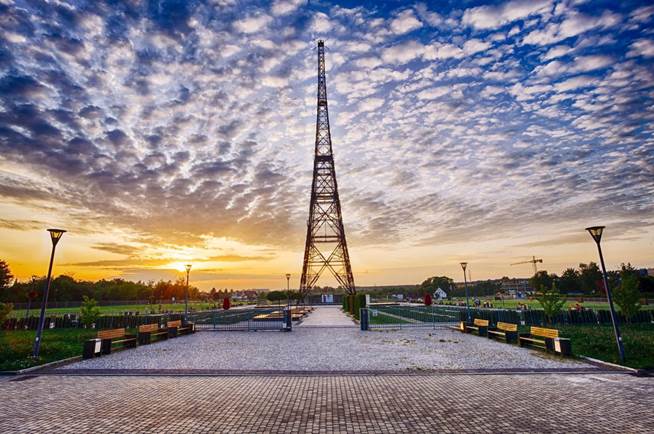 Na zdjęciu widzimy malowniczy widok placu z Radiostacją Gliwice (najwyższą drewnianą konstrukcję na świecie) w tle. Wieża, wznosząca się ku niebu, jest centralnym punktem kompozycji. Na pierwszym planie widać dojscie do wieży z kostki brukowej, wzdłuż niego umieszczone są ławki. Całość zdjęcia dopełnia piękne, kolorowe niebo z chmurami o zachodzie słońca. 