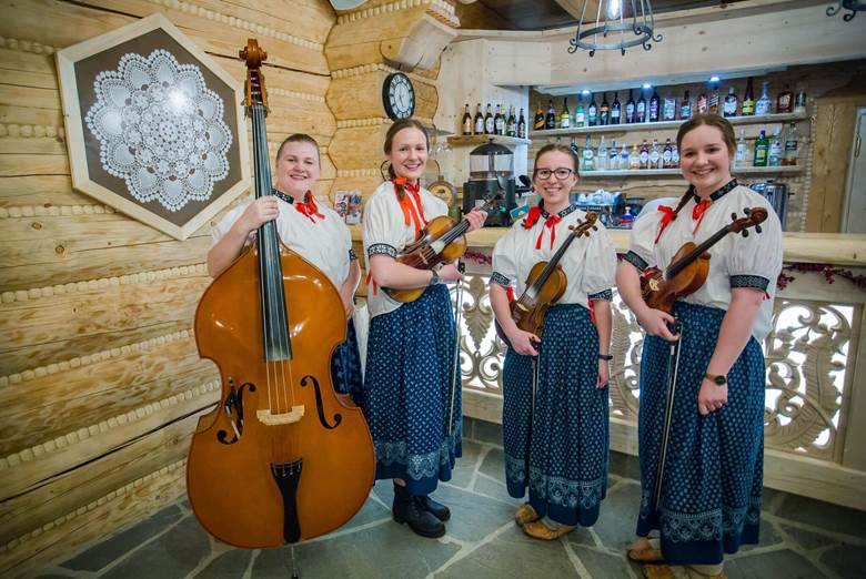 Kapela góralska z Istebnej: 4 góralki w białogranatowych strojach ludowych. Trzy z nich trzymaja w rękach skrzypce, a czwarta kontrabas. W tle wnętrze drewnianej karczmy.